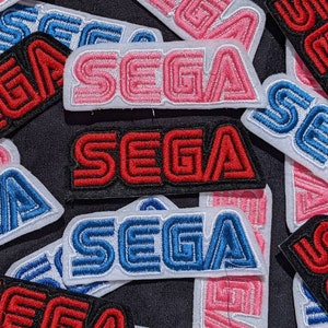 Könnte beinhalten: Eine Sammlung von gestickten Patches mit dem Wort "SEGA" in verschiedenen Farben und Stilen. Die Patches sind aus Stoff und haben einen weißen Hintergrund mit schwarzen Umrissen. Die Patches sind perfekt, um Kleidung, Taschen oder anderen Accessoires einen Retro-Gaming-Touch zu verleihen.