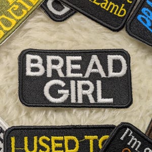 May include: Black embroidered patch with white text that reads "BREAD GIRL".