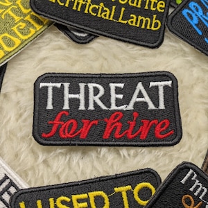 May include: Black embroidered patch with white text that reads "THREAT for hire" in red lettering.