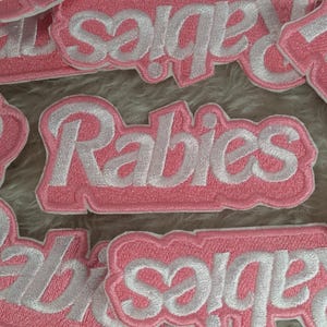 Op de afbeelding: Roze stoffen patches met witte geborduurde tekst. De patches lezen "Rabies" en "Saligia".