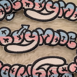 May include: Embroidered patches spelling "Baby Mode" in a bubble letter style. The patches use pink, light blue, and white, with a black outline. The patches are displayed on a fluffy, light-coloured surface.