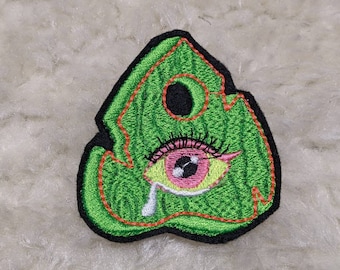 Puede incluir: Parche bordado verde con un ojo grande en el centro. El ojo es rosa y blanco con pestañas negras. El parche tiene forma de hoja.