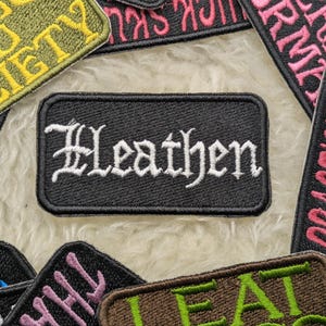 May include: A black embroidered patch with white lettering that reads "Heathen". The patch is surrounded by other colourful embroidered patches with various words and designs.