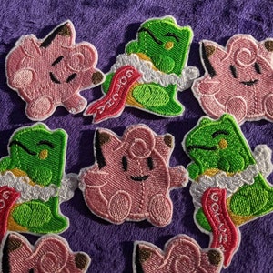 May include: Eight embroidered iron-on patches featuring pink and green cartoon characters. The patches have white trim and red ribbons with the text "Gotcha" and "Good Luck".