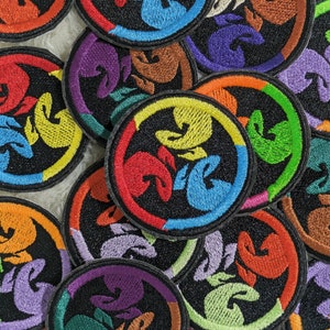 Könnte beinhalten: Eine Sammlung bunter gestickter Patches mit einem stilisierten Design aus drei ineinandergreifenden Figuren. Die Patches sind rund und haben einen schwarzen Hintergrund mit einem Regenbogen an Farben.