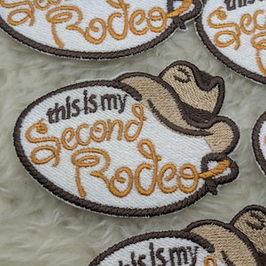 Peut inclure: Patch thermocollant brodé blanc avec des points de couture marrons. Le patch représente un chapeau de cowboy et le texte "This is my Second Rodeo".