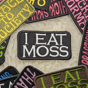 May include: A collection of colorful embroidered patches with various phrases, including "I Eat Moss". The patches are made of fabric and have a black, white, and green color scheme.