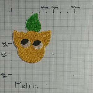 May include: A yellow embroidered patch with a green leaf on top. The patch has two large white eyes and a small mouth. The patch is approximately 6 cm wide and 6 cm tall.