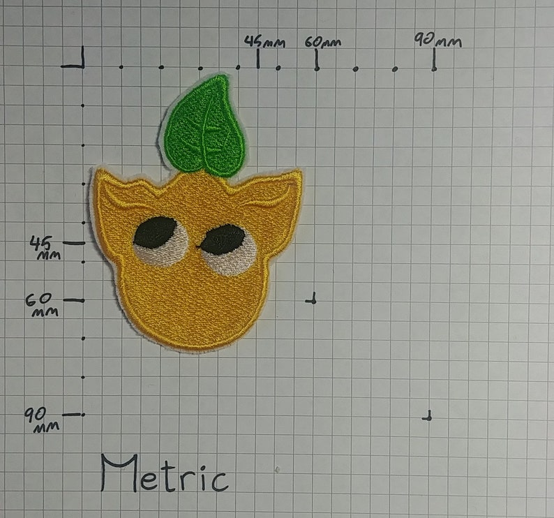 May include: A yellow embroidered patch with a green leaf on top. The patch has two large white eyes and a small mouth. The patch is approximately 6 cm wide and 6 cm tall.