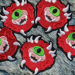 Puede incluir: Parches bordados con un diseño circular rojo, un ojo verde, dientes blancos y bordes dentados. Los parches tienen un borde negro y están dispuestos sobre una superficie texturizada azul y gris. Estos parches son adecuados para chaquetas, bolsos u otros artículos de tela.