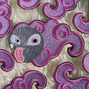 May include: Purple and pink embroidered patches shaped like clouds with a black and white cartoon character with a pink tongue sticking out.