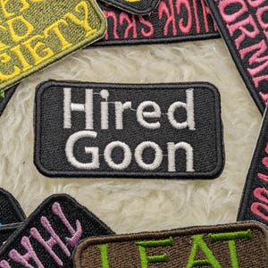Può includere: Toppa ricamata in bianco e nero con il testo "Hired Goon" in lettere bianche. La toppa è circondata da altre toppe colorate con diversi testi.