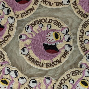 May include: A circular embroidered patch featuring a light purple, multi-eyed monster on a cream background. The monster has a large central eye with a yellow iris and multiple smaller eyes on stalks. The text BEHOLD 'ER? HARDLY KNOW 'ER is stitched.