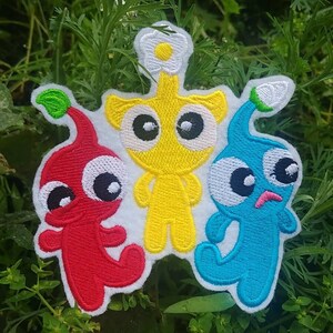 May include: Embroidered patch featuring three cartoon characters. The characters are red, yellow, and blue. The red character has a green leaf on its head. The yellow character has a white flower on its head. The blue character has a blue leaf on its head.