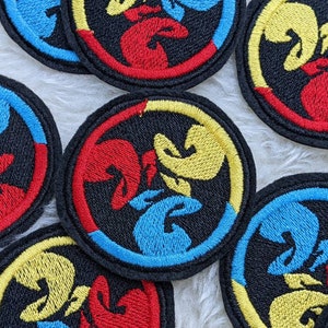 May include: A set of six embroidered patches featuring a stylized design of three figures in a circle. The figures are outlined in black and filled with red, yellow, and blue.