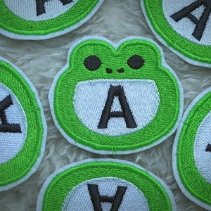 May include: Five green embroidered patches with white centers and black letter 'A's. One patch features a cartoon frog with a white center and black letter 'A'.