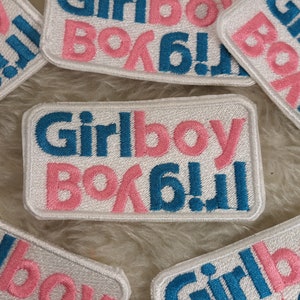 May include: Four white patches with embroidered text in pink and blue. The text reads "Girlboy" with the "Girl" in blue and the "Boy" in pink. The patches are on a fluffy white surface.