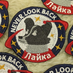 May include: A set of five embroidered patches featuring a cartoon dog with the text "Never Look Back" and "Laika" in red and white lettering. The patches are on a white fluffy surface.