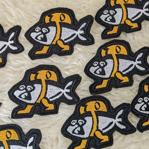 Op de afbeelding: Een set van tien strijkbare patches met een cartoonvis met een gele vin en een wit lichaam. De patches zijn zwart met witte en gele borduursels.
