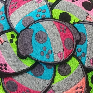 May include: A collection of embroidered patches, each depicting a colourful game controller design. The patches are edged in black and feature pink, blue, green, and grey sections. Each patch has a cartoon face.