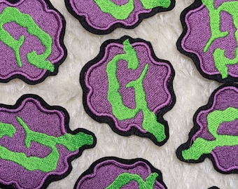 May include: A set of six embroidered iron-on patches featuring a stylized letter G in a green and purple color scheme. The patches are shaped like a rounded rectangle with a scalloped edge.