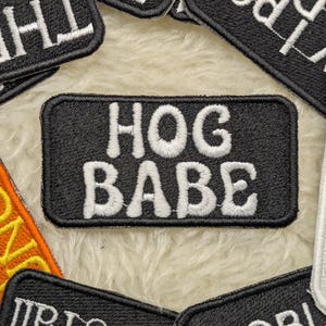 Puede incluir: Parche rectangular negro con las palabras "HOG BABE" bordadas en blanco. También se ven otros parches con texto blanco sobre fondos negros y un parche naranja con texto amarillo.