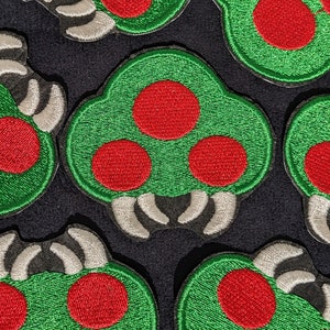 Puede incluir: Cinco parches bordados con una criatura verde de tres ojos y patas blancas. La criatura tiene tres ojos rojos y un cuerpo verde.