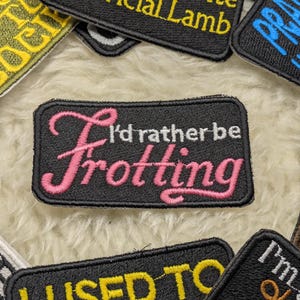 Op de afbeelding: Zwarte geborduurde patch met witte en roze letters met de tekst "I'd rather be frottering".