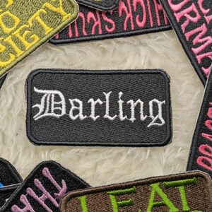Peut inclure: Un patch brodé noir avec des lettres blanches qui disent "Darling". Le patch est entouré d'autres patchs brodés colorés avec différents mots et motifs.