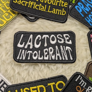 Könnte beinhalten: Ein schwarzer und weißer gestickter Aufnäher mit dem Text "LACTOSE INTOLERANT" in weißen Buchstaben. Der Aufnäher ist von anderen bunten Aufnähern mit verschiedenen Texten umgeben.