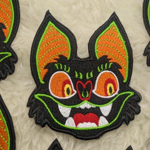 Op de afbeelding: Een geborduurde patch met een cartoon-vleermuis met oranje ogen en een brede, tandenrijke grijns. De vleermuis heeft grote, oranje en groene oren.