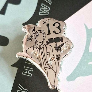 May include: A silver-toned metal pin featuring an engraved outline of a person in a suit, the number 13, and the text JIMIN. The design includes small floral accents and has an irregular, organic shape with a polished, reflective surface finish.