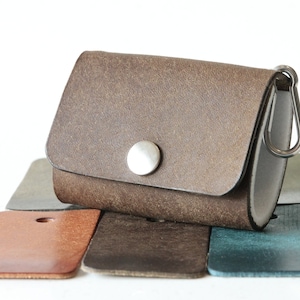 Könnte beinhalten: Eine kleine, braune Ledertasche mit einem silbernen Druckknopfverschluss und einem Metallclip. Die Tasche liegt auf einer Sammlung von Lederfarbmustern in verschiedenen Farben, darunter Braun, Grau und Türkis. Die Tasche ist etwa 7.6 cm breit.