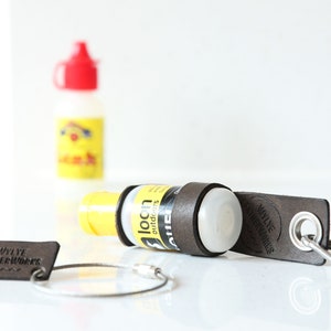 Könnte beinhalten: Ein brauner Leder-Schlüsselanhänger mit dem Text "WYLIE LEATHERWORKS" und einem silbernen Schlüsselring. Im Hintergrund eine gelb-weiße Flasche mit rotem Deckel. Ein braunes Lederband umschließt die Flasche.