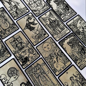 May include: A set of tarot cards with black and white illustrations on a silver background. The cards are arranged in a random pattern. The cards include The Magician, The Hierophant, The Chariot, The Fool, The Sun, Wheel of Fortune, Justice, Queen of Pentacles, and High Priestess.