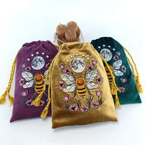May include: Three velvet drawstring pouches in purple, gold, and dark green, each featuring an embroidered design of a bee, pink flowers, a moon, and stars. The pouches have gold cord drawstrings and are arranged with a small bundle of beeswax candles.