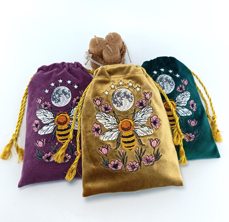 May include: Three velvet drawstring pouches in purple, gold, and dark green, each featuring an embroidered design of a bee, pink flowers, a moon, and stars. The pouches have gold cord drawstrings and are arranged with a small bundle of beeswax candles.