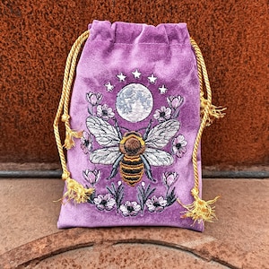 May include: A purple velvet drawstring bag with an embroidered design. The design features a bee with outstretched wings, a full moon, stars, and flowers. The bag has gold-colored drawstrings.