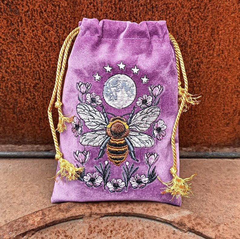 May include: A purple velvet drawstring bag with an embroidered design. The design features a bee with outstretched wings, a full moon, stars, and flowers. The bag has gold-colored drawstrings.