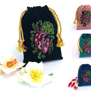 May include: A collection of drawstring pouches in black, pink, teal, and blue, each featuring an embroidered anatomical heart surrounded by green monstera leaves. The pouches have gold-colored braided cords and are displayed with white flowers.