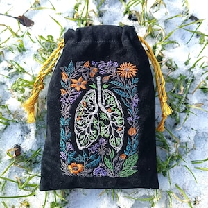 Puede incluir: Bolsa negra con cordón con un diseño bordado de pulmones rodeados de flores y follaje coloridos. El bordado utiliza hilo naranja, morado, verde y blanco. El cordón es de color amarillo dorado.
