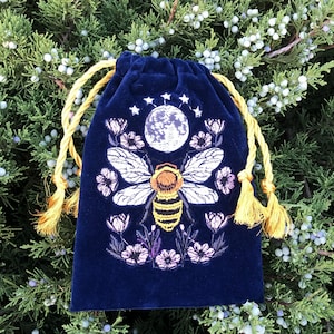 May include: A deep blue velvet drawstring pouch featuring an embroidered design of a central bee with yellow and black stripes, white wings, a full moon above, small stars, and surrounding pink and purple flowers with golden yellow tassels.