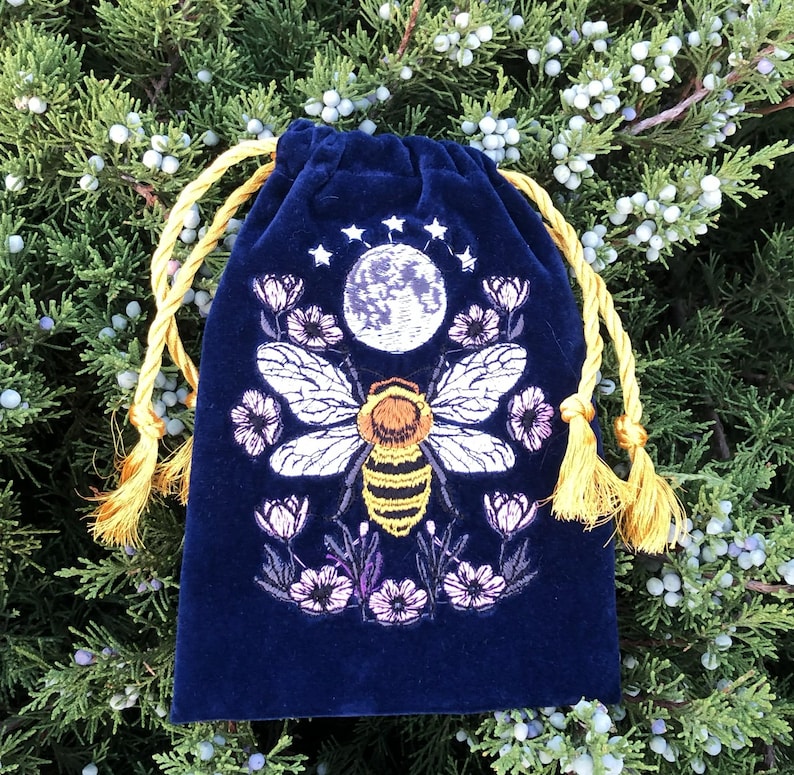May include: A deep blue velvet drawstring pouch featuring an embroidered design of a central bee with yellow and black stripes, white wings, a full moon above, small stars, and surrounding pink and purple flowers with golden yellow tassels.