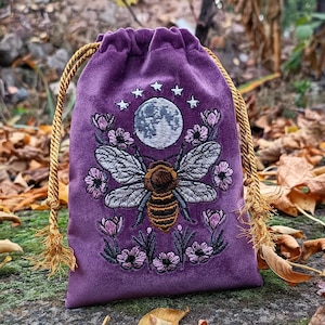 May include: A purple velvet drawstring bag with an embroidered design. The design features a bee with detailed wings, a full moon, stars, and floral accents. The bag has a gold rope drawstring closure and tassel ends.