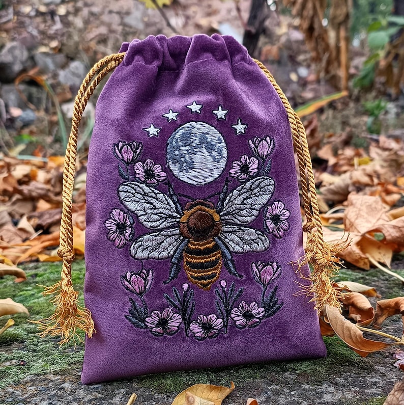 May include: A purple velvet drawstring bag with an embroidered design. The design features a bee with detailed wings, a full moon, stars, and floral accents. The bag has a gold rope drawstring closure and tassel ends.