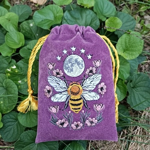 May include: A purple velvet drawstring pouch featuring an embroidered design of a central honeybee with yellow and black stripes, surrounded by pink flowers and a grey moon topped with five white stars, finished with yellow braided cord ties.
