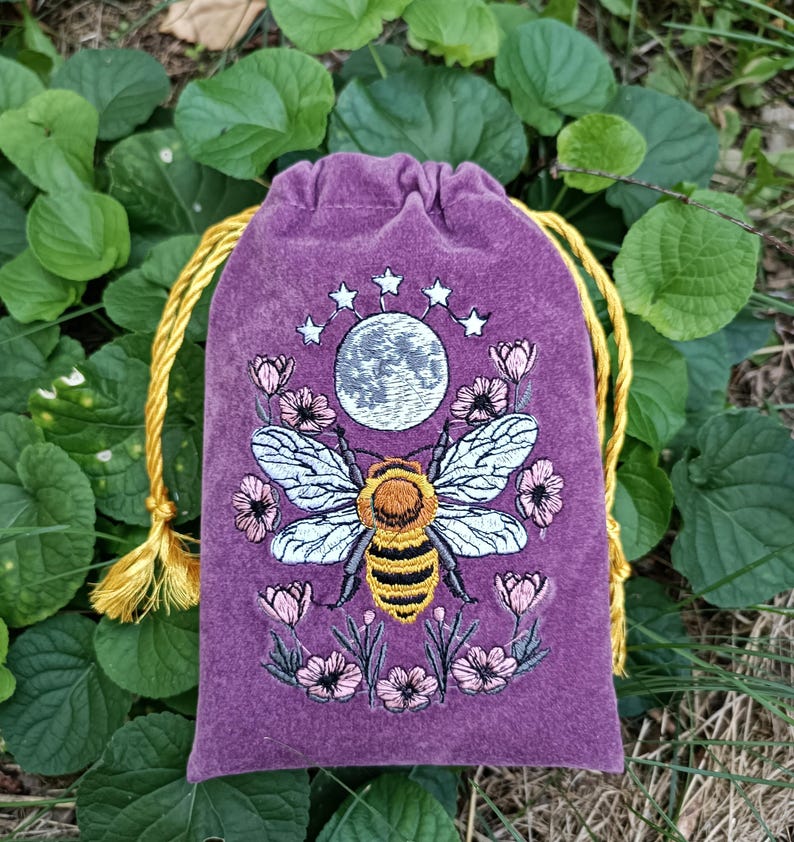 May include: A purple velvet drawstring pouch featuring an embroidered design of a central honeybee with yellow and black stripes, surrounded by pink flowers and a grey moon topped with five white stars, finished with yellow braided cord ties.