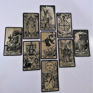 May include: A set of ten tarot cards with black and white illustrations on a gold background. The cards include The Magician, The Hierophant, The Chariot, The Fool, The Sun, Wheel of Fortune, Justice, Queen of Cups, Queen of Pentacles.