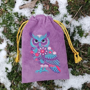 Op de afbeelding: Een paarse trekkoordtas met een geborduurd uilenontwerp. De uil heeft turquoise, roze en paarse details, met een bloemenpatroon op de borst. De tas heeft gouden trekkoorden en kwastjes, tegen een achtergrond van sneeuw en gras.