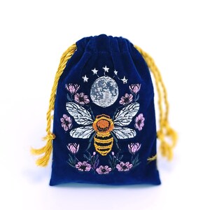 May include: A deep blue velvet drawstring pouch featuring an embroidered design of a central bee with yellow and black stripes, white wings, a full moon above, small stars, and pink flowers surrounding the bee on the front surface.
